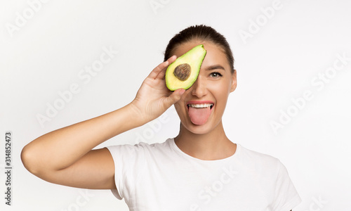 Woman Holding Avocado Half Covering Eye, Showing Tongue, White Background