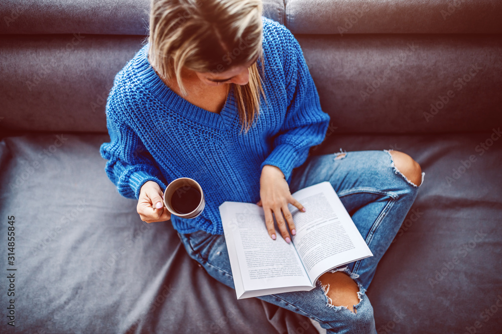 Top view of blond caucasian young woman in sweater sitting on sofa in living room, drinking coffee and reading book. - obrazy, fototapety, plakaty 