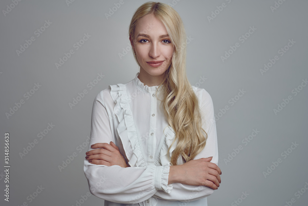 Young and beautiful long-haired blond woman in white blouse standing with her arms folded and looking at camera with a smirk. Front half-length portrait against grey background Young and beautiful long-haired blond woman in white blouse standing with her arms folded and looking at camera with a smirk. Front half-length portrait against grey background