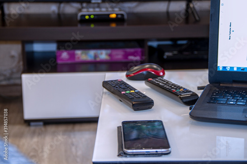 Two TV remotes in the background of a room with a laptop and a smartphone with a TV set-top box.