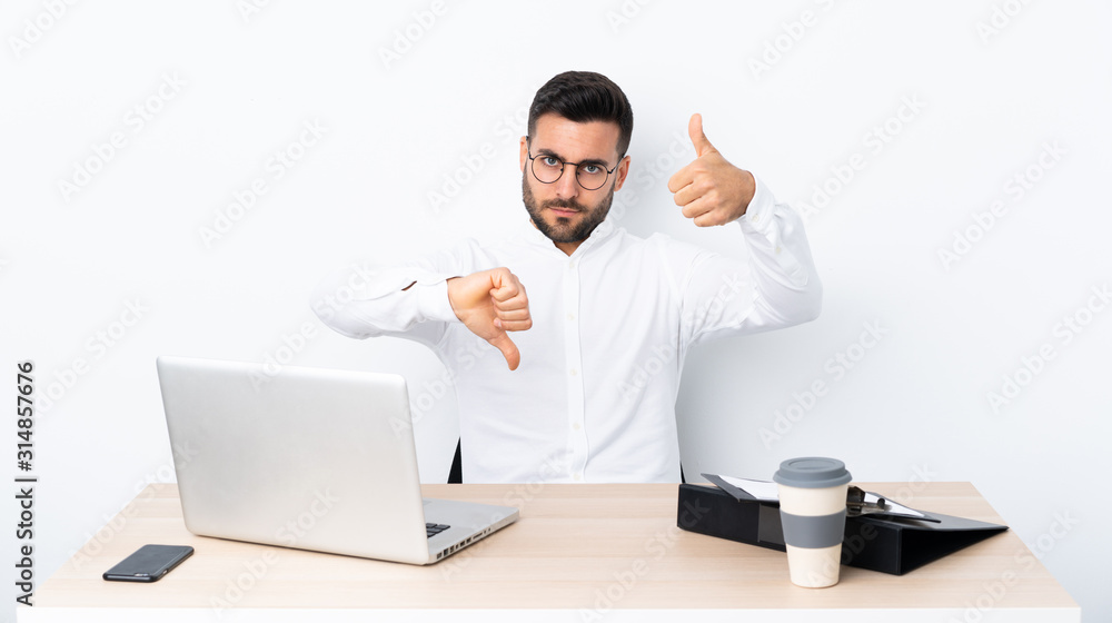 Young businessman in a workplace making good-bad sign. Undecided between yes or not Young businessman in a workplace making good-bad sign. Undecided between yes or not