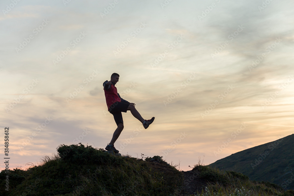 Dark silhouette of a hiker balancing on a summit stone in evening mountains.