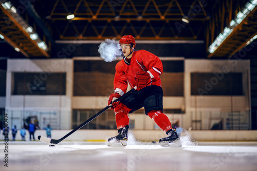 Dedicated strong hockey player in red uniform with protective helmet on head skating and going to goal with stick and puck.