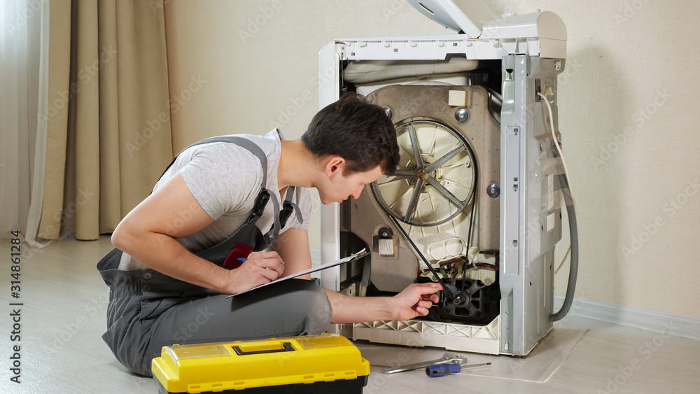 Obraz premium young serviceman checks broken washing machine loader with open back side panel on floor in room closeup