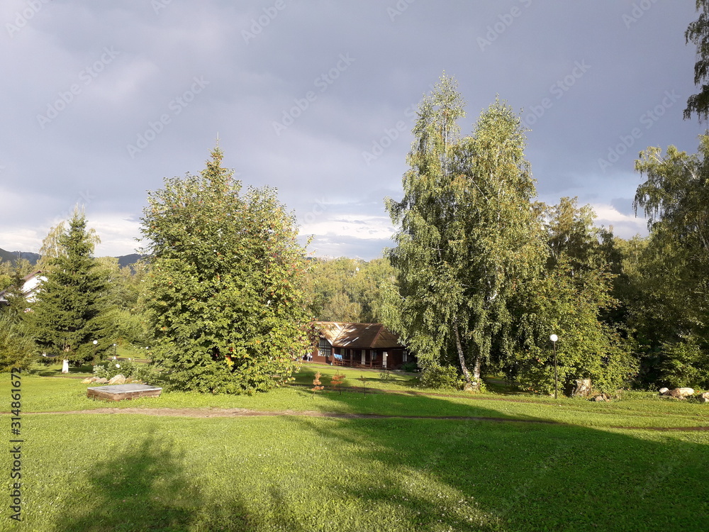 Green glade illuminated by the evening sun. Wooden house. Shadows from the trees. Green glade illuminated by the evening sun. Wooden house. Shadows from the trees.
