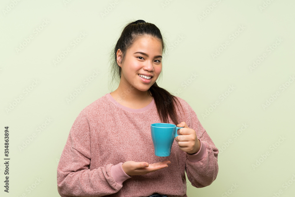 Young teenager Asian girl over isolated green background holding hot cup of coffee Young teenager Asian girl over isolated green background holding hot cup of coffee