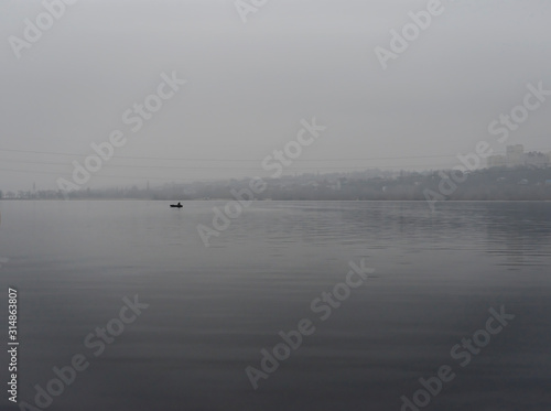 Fishermen on an inflatable boat in the middle of the river against the background of the city's high-rise buildings. Foggy weather, weekend, hobby.