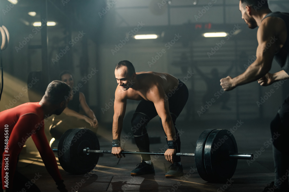 Smiling athletic man lifting barbell with support of his friends in a gym. Smiling athletic man lifting barbell with support of his friends in a gym.