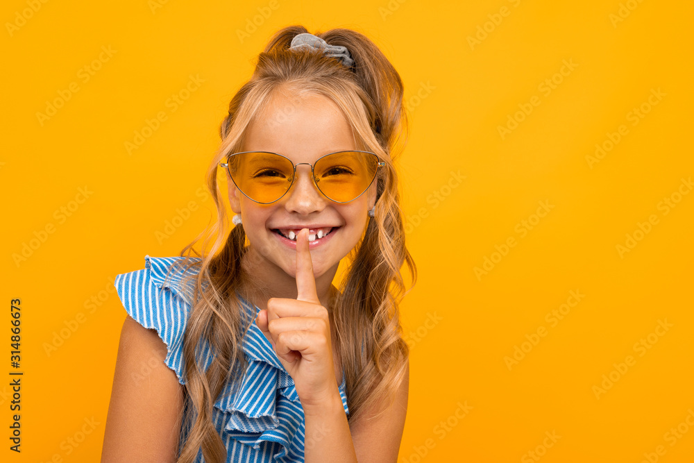 charming blonde girl in a dress and sunglasses asks to be quieter, holds a finger at her mouth on a yellow background with copy space charming blonde girl in a dress and sunglasses asks to be quieter, holds a finger at her mouth on a yellow background with copy space