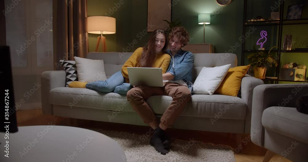 Young Caucasian happy in love couple resting on the couch and hugging while watching something on laptop computer together late at night and smiling. Indoors. Young Caucasian happy in love couple resting on the couch and hugging while watching something on laptop computer together late at night and smiling. Indoors.
