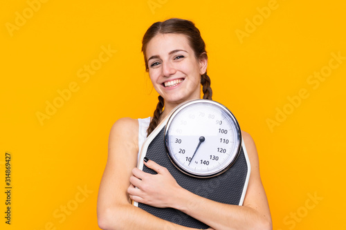 Young woman over isolated yellow background with weighing machine