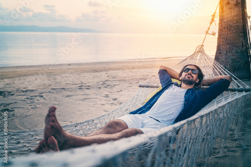 Young handsome Latin man in sunglasses relaxing in a hammock on the beach at sunset on the beach. photo