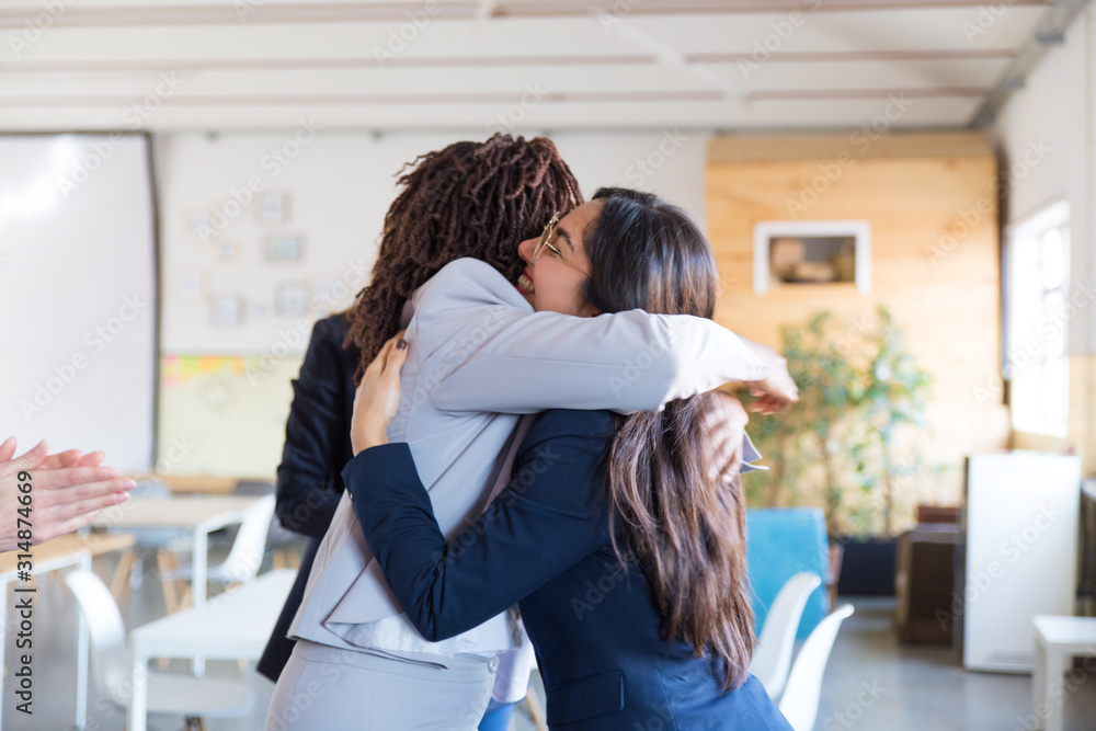 Happy businesswomen hugging in office. Side view of cheerful multiethnic young businesswomen meeting each other in office. Business concept Happy businesswomen hugging in office. Side view of cheerful multiethnic young businesswomen meeting each other in office. Business concept