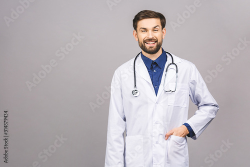 Portrait of cheerful smiling young doctor with stethoscope over neck in medical coat standing against isolated gray background.