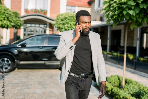 African american businessman in a suit speaking on smartphone, walking outdoors on the background of his black car and office buildings. photo