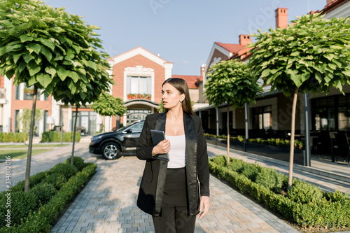 Portrait of young Caucasian pretty concentrated businesswoman walking with digital tablet against blcak car and modern office center outdoors