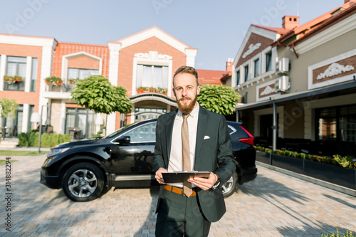Young businessman or sales manager in dark suit standing outdoor on the yard of business center with new black car on background