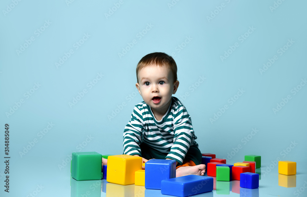 Little baby boy in stylish casual clothing barefoot sitting on floor and playing with colorful toy cubes over blue wall background Little baby boy in stylish casual clothing barefoot sitting on floor and playing with colorful toy cubes over blue wall background