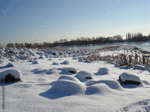 Schneebedecktes Flussufer als winterliches Idyll und Winterlandschaft lädt zum Winterspaziergang ein und zeigt Spuren im Schnee und Raureif mit Schneekristallen © sunakri