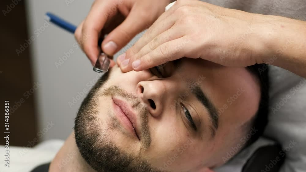 A client has a beard. He has dark hair. A barber is working in salon. A client has a beard. He has dark hair. A barber is working in salon.