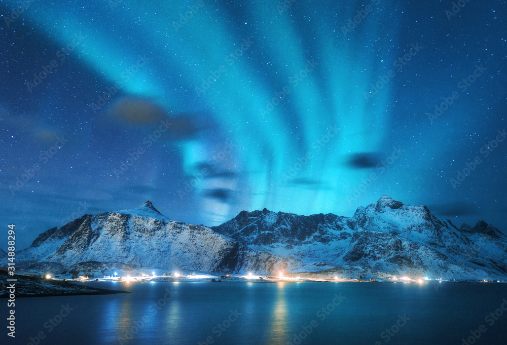 Aurora borealis over sea, snowy mountains and city lights at Northern lights in Lofoten islands, Norway. Blue starry sky with polar Winter landscape with reflected in water Stock