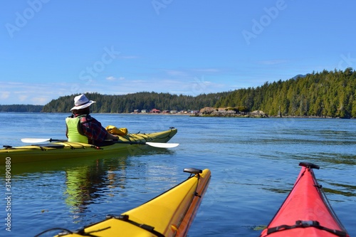 Group of friends on sea kayak in Pacific Ocean near Vancouver Island. Colorful kayaks, trees on the island, man with west, hat and paddle. Blue sky. 