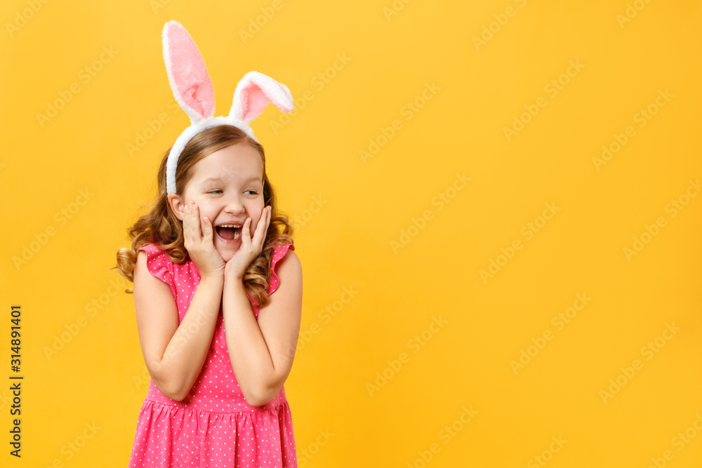 Funny little girl in Easter bunny ears on a yellow background. The child opened his mouth in surprise and looks at the empty space Funny little girl in Easter bunny ears on a yellow background. The child opened his mouth in surprise and looks at the empty space