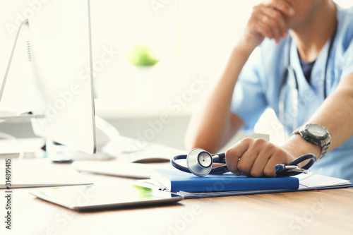 Fototapeta Naklejka Na Ścianę i Meble -  Serious male doctor looking at computer monitor while holding stethoscope in one hand and propped his head with other hand.