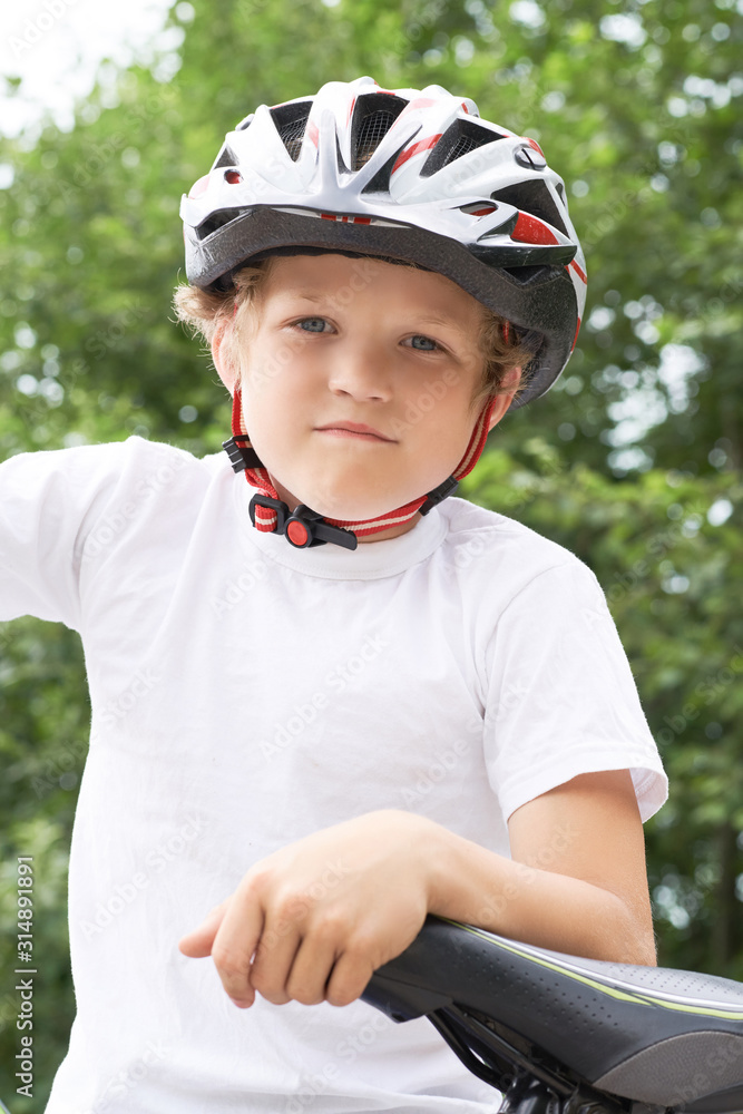 Little Caucasian boy in protective helmet stands leaning on the bike posing for the camera. Teenager ready to ride bicycle in park on summer day. Weekend activity. Little Caucasian boy in protective helmet stands leaning on the bike posing for the camera. Teenager ready to ride bicycle in park on summer day. Weekend activity.