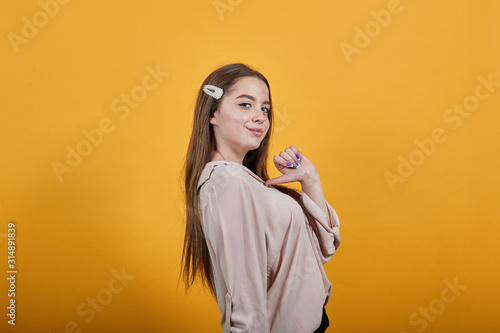 Attractive caucasian young woman staying sideways, pointing thumb at herself wearing fashion pastel shirt isolated on orange background in studio. People sincere emotions, lifestyle concept.
