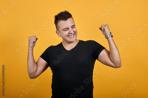 Charming young man wearing black shirt isolated on orange background in studio doing winner gesture, smiling, keeping fists up. People sincere emotions, lifestyle concept.