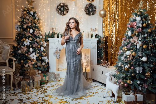 Beautiful young woman with glass of champagne over Christmas background. Girl with a bright make-up, celebrates Christmas,New Year. Beautiful model woman in dress posing in luxurious interior