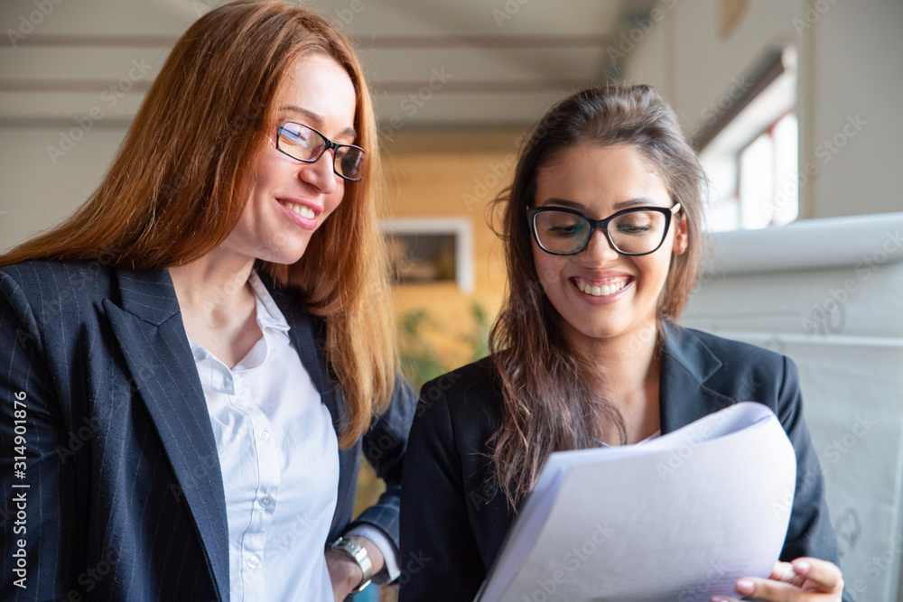 Fototapeta premium Two smiling women studying new project. Front view of cheerful colleagues reading paper document. Teamwork concept
