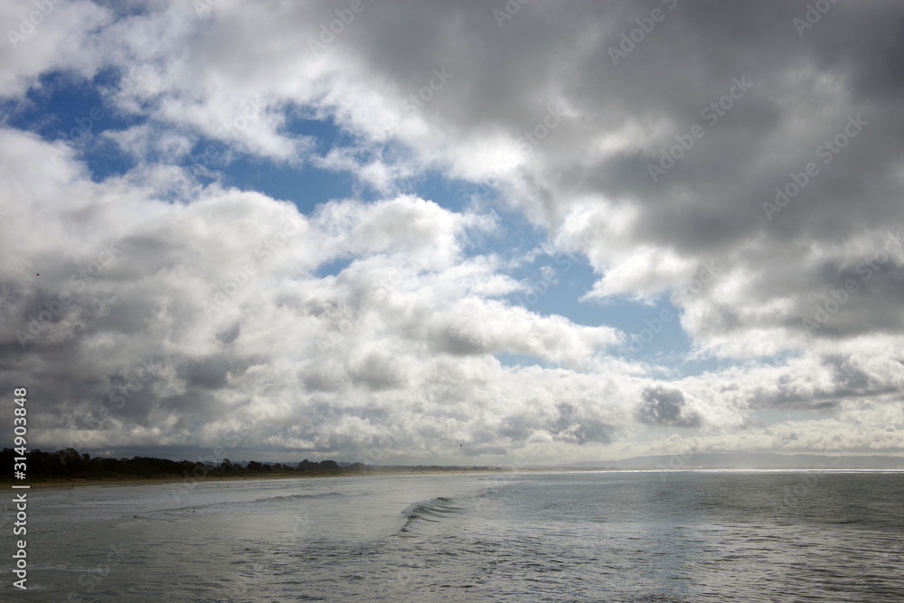 Panoramic view of heavy storm clouds with some blue sky patches over a wide beach and ocean waters on a California winter day