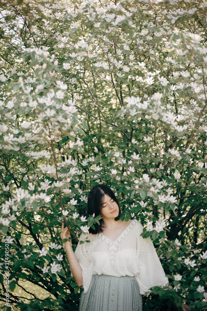 Stylish boho woman posing in white flowers in spring park. Hipster girl in boho outfit standing in white blooms of a tree. Calm portrait of beautiful brunette woman in spring Stylish boho woman posing in white flowers in spring park. Hipster girl in boho outfit standing in white blooms of a tree. Calm portrait of beautiful brunette woman in spring