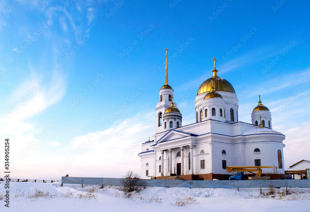 White beautiful Orthodox Church under construction in winter time. Snowy panorama of blue sky and christian church with golden domes.