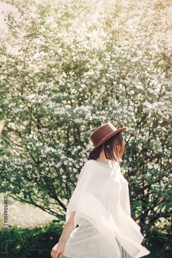 Happy boho woman in hat dancing in sunny light near blooming tree with white flowers in spring park. Stylish hipster girl enjoying spring and moving, having fun. Atmospheric moment