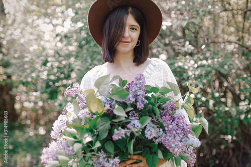 Stylish boho woman in hat holding lilac flowers bouquet in sunny spring park. Calm portrait of beautiful hipster girl standing with purple lilac in spring garden. Copy space