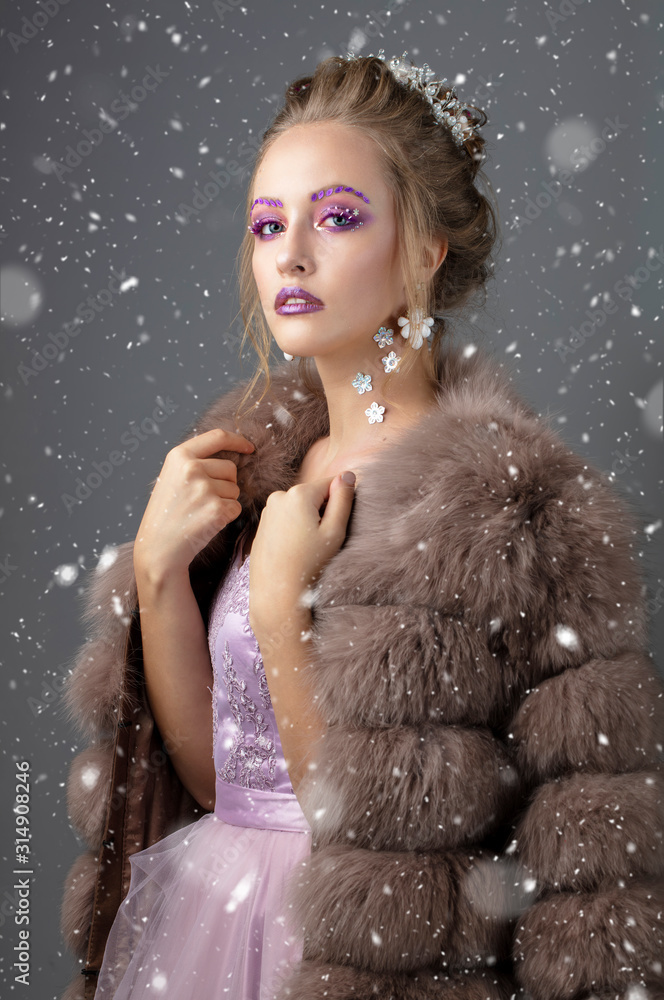 Beautiful model with fashionable make-up in a fur coat. Luxury winter girl on a gray background strewn with snow.