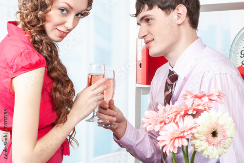 A young man, an employee of the company shows courtship to his colleague-a young woman with curly hair. The man gave flowers and offers to drink champagne in the office on Valentine's day. Flirting, 