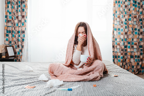 The concept of diseases. A woman sits on the bed covered with a blanket, and blows her nose in a handkerchief. Next to her are pills, a thermometer and handkerchiefs. Window in the background photo