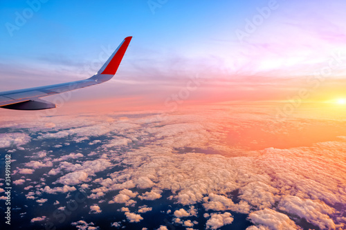 modern airplane wing view of orange sunset sky landscape against beautiful red cloudscape background. Wide outside view of passenger aircraft window. Plane flying over sea with scenic sunrise clouds