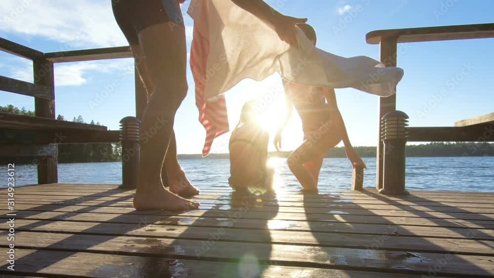 A man wraps a towel around his wife and daughter after swimming in the lake sitting on a wooden jetty at sunset after the sauna in summer white night in Finland.