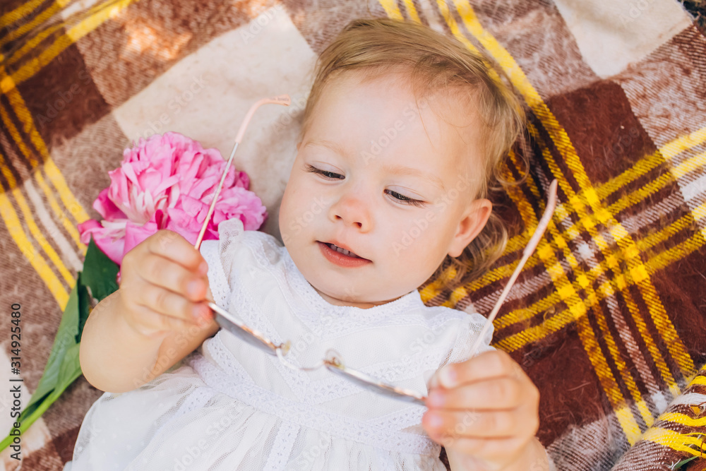 Little cute baby in a white dress with a pink peony in hands lie on a plaid in the park. Family outdoor recreation. Family summer picnic in the park. Girl outdoors surrounded by pink peonies. Little cute baby in a white dress with a pink peony in hands lie on a plaid in the park. Family outdoor recreation. Family summer picnic in the park. Girl outdoors surrounded by pink peonies.