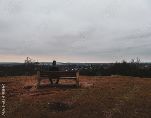 One man sitting alone on a bench on top of a hill overlooking flat countryside. Taken on a dark, overcast Autumn day in Sandy, Bedfordshire.