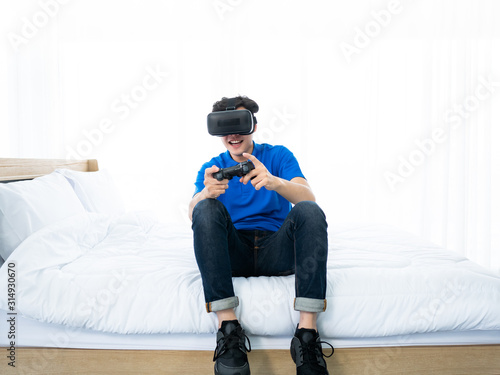 20s Asian young man fun and enjoy from playing virtual reality headset with joystick controller digital technology device for simulation futuristic visual world in glasses with friends in his bedroom © MergeIdea