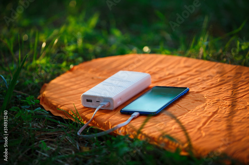 Using a power Bank to charge your phone and gadgets in the field. White portable battery charges on a backpack, on a stump, on a tourist Mat.