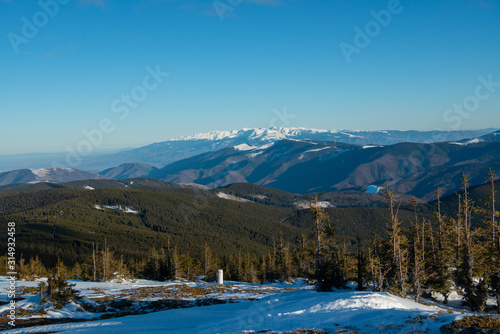 A winter landscape at midday from the peak of a mountain with some snowy mountain peaks in the background and a a slope with trees in the foreground