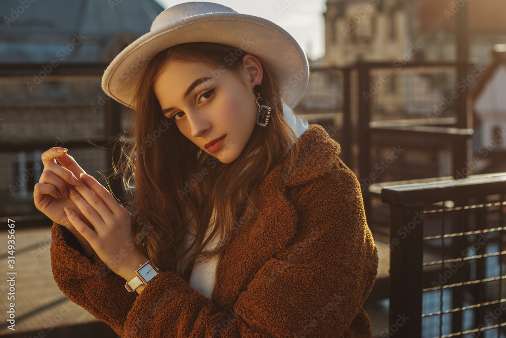 Outdoor fashion portrait of young elegant fashionable brunette woman, model wearing stylish white hat, wrist watch,  brown faux fur coat, posing at sunset, in European city. Copy empty space for text - obrazy, fototapety, plakaty 