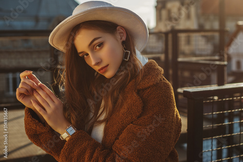 Outdoor fashion portrait of young elegant fashionable brunette woman, model wearing stylish white hat, wrist watch,  brown faux fur coat, posing at sunset, in European city. Copy empty space for text photo